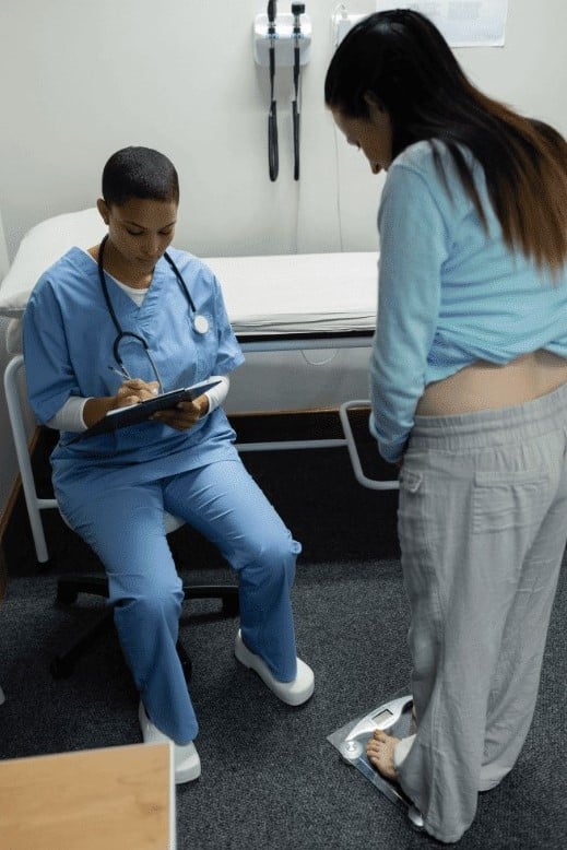 Black health care provider with clipboard writing down a woman's weight as she stands on scale